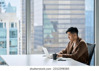 Young adult serious busy Asian business man manager wearing suit typing on laptop using computer working on digital project financial market data software management sitting at desk in office.