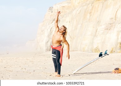 Young adult shirtless surfer with dreadlocks wearing wetsuit warming up on beach preparing for summer evening surfing session - stretching routine for surfing and active lifestyle concept, Portugal - Powered by Shutterstock