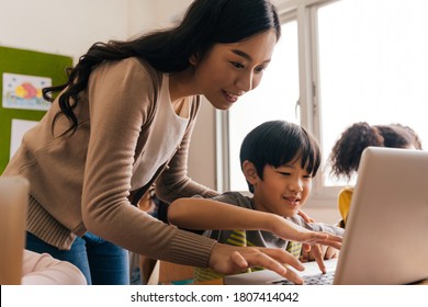 Young Adult Smiling Beautiful Asian Teacher Helping Elementary Student Boy With Laptop In Computer Classroom. Information Technology Class In Primary School Concept.