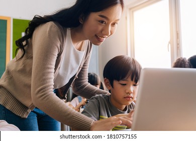 Young Adult Smiling Beautiful Asian Teacher Helping Elementary Student Boy With Laptop In Computer Classroom. Information Technology Class In Primary School Concept.