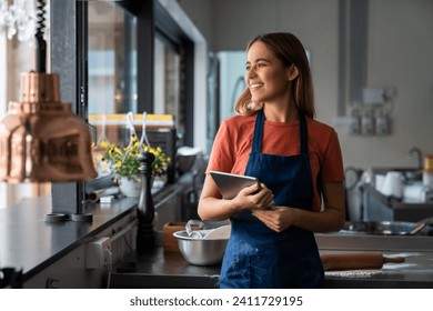 Young adult smiling female baker looking through window while standing in pastry kitchen and holding digital tablet.