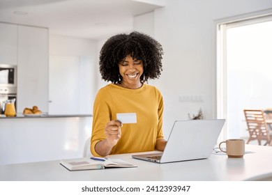 Young adult smiling happy African American woman holding credit card using laptop computer paying or buying online making ecommerce shopping purchase sitting at home table in kitchen. - Powered by Shutterstock