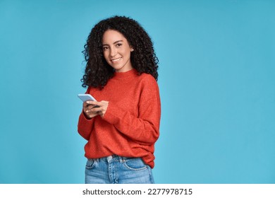 Young adult smiling happy pretty latin woman holding mobile phone device, doing ecommerce online shopping on cell, using apps on cellphone standing isolated on blue background. - Shutterstock ID 2277978715