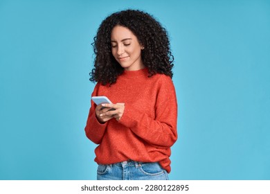 Young adult smiling happy pretty latin woman holding mobile phone looking at smartphone, typing message doing ecommerce shopping on cell, using trendy apps on cellphone isolated on blue background.