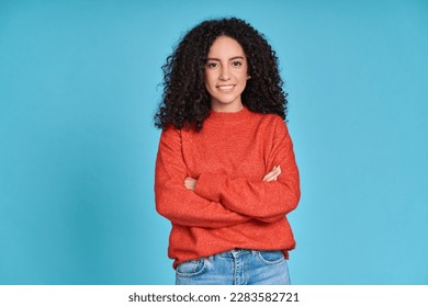 Young adult smiling latin professional woman, happy pretty curly hispanic female model student wearing orange sweater standing looking at camera arms crossed isolated on blue background, portrait.