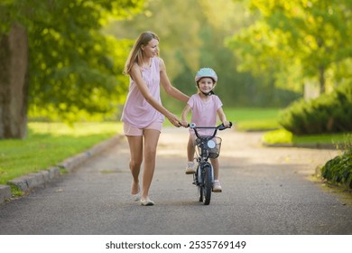 Young adult smiling mother teaching happy beautiful little girl to ride on first bike on sidewalk at city park. Learning to keep balance. Warm summer day. Cute 5 years old toddler. Front view. - Powered by Shutterstock