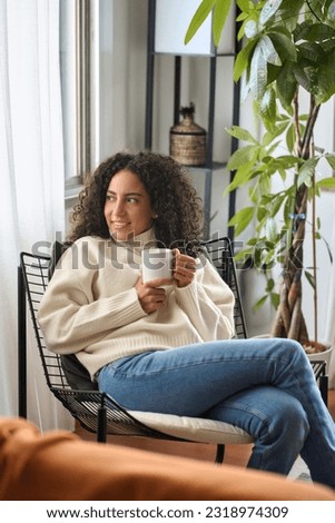 Young adult smiling pretty latin woman sitting on chair holding cup drinking tea or coffee relaxing at home. Happy calm lady enjoying warm hot drink with mug in hands daydreaming. Vertical