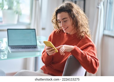 Young adult smiling pretty woman sitting on chair holding smartphone using cellphone modern technology, looking at mobile phone while remote working or learning, texting messages at home office.