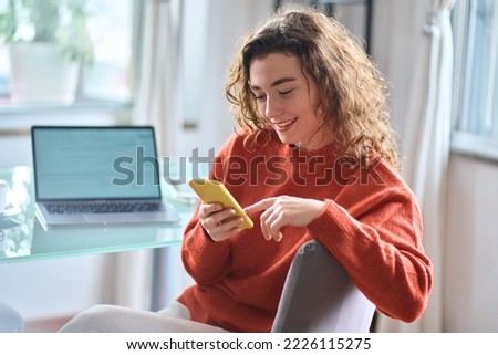 Young adult smiling pretty woman sitting on chair holding smartphone using cellphone modern technology, looking at mobile phone while remote working or learning, texting messages at home office.