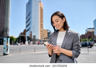 Young adult smiling professional Asian business woman wearing suit holding smartphone looking at mobile phone using apps on cellphone technology texting standing on urban city street outside. - Shutterstock ID 2217353233