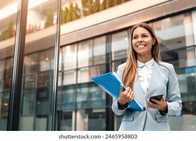 Young adult smiling professional business woman wearing suit holding smartphone and folder with files while walking in urban city environment. - Powered by Shutterstock