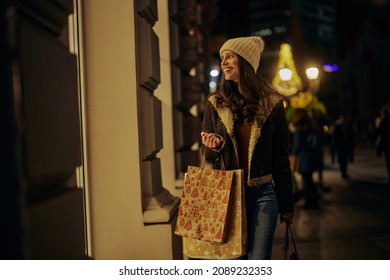 Young Adult Smiling Woman Walking On Decorated City Streets With Christmas Lights, Carrying Shopping Bags, Looking At The Shop Window, Searching More Gifts For Friends And Family