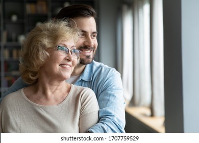 Young Adult Son Hugs Elderly Mother, Family Smiling Looking Out The Window Standing In Living Room At Home. Offspring And Future, Grown-up Child Give Care To Mom, Visit Her Provide Attention Concept