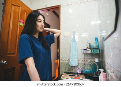 Young Adult Southeast Asian Woman Practice Self And Checking By Mirror In Bathroom At Home. Mental Health Maintain Lifestyle After Wake Up Concept. Beautiful Face With Long Hair Wear Blue Shirt.