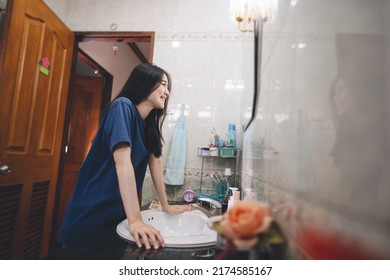 Young Adult Southeast Asian Woman Practice Self And Checking By Mirror In Bathroom At Home. Mental Health Maintain Lifestyle After Wake Up Concept. Beautiful Face With Long Hair Wear Blue Shirt.