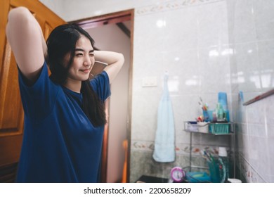 Young Adult Southeast Asian Woman Practice Self And Checking By Mirror In Bathroom At Home. Mental Health Maintain Lifestyle After Wake Up Concept. Beautiful Face With Long Hair Wear Blue Shirt.