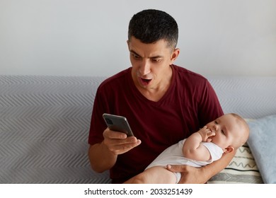 Young Adult Surprised Man Wearing Casual Style Maroon T Shirt Holding Baby Girl Or Boy In Arms And Using Mobile Phone While Sitting On Sofa In Living Room, Get Shocking News From Email.