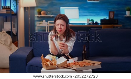 Young adult using video call remote technology sitting on living room sofa. Woman talking on online conference while eating fries and fast food. Person with internet and takeaway food