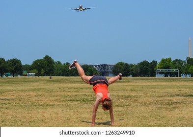 Young Adult Woman Attempts To Do A Cartwheel In A Park, But Fails. Airplane And Washington Monument In The Background.