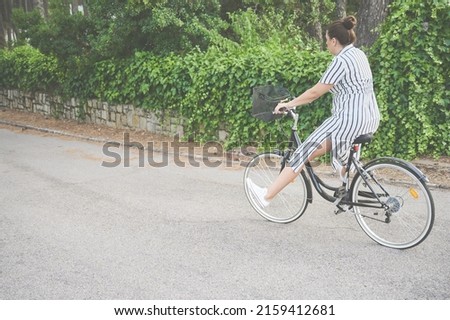 Young adult woman with a bow in her hair and a striped dress enjoys a beautiful summer day riding a bike. She rides down a street with her feet off the pedals. Summertime concept.