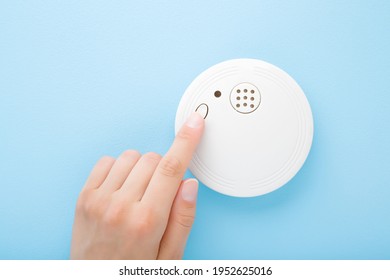 Young Adult Woman Finger Pushing Button Of New White Plastic Smoke Alarm. Light Blue Table Background. Pastel Color. Closeup. Safety Concept.
