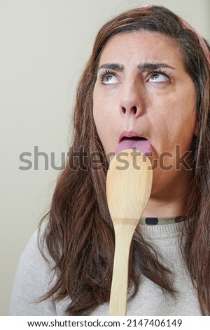 A young adult woman gesticulates as she tastes a new recipe using a wooden cookware spoon. She is licking the spoon looking up.