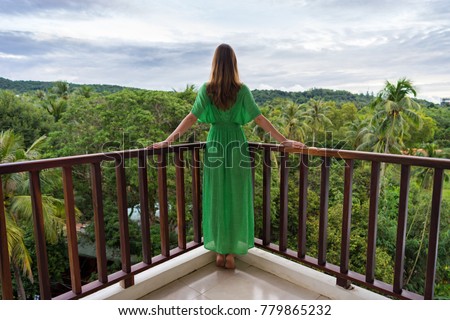 Young adult woman in green dress is standing on balcony in tropical country in luxury hotel and look forward on greenery jungle. Back view