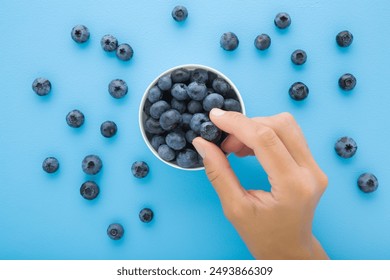 Young adult woman hand fingers taking fresh blueberries from white bowl on light blue table background. Pastel color. Eating healthy berries. Closeup. Top view. - Powered by Shutterstock