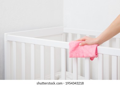Young adult woman hand holding pink dry rag and wiping edge of white crib in bedroom. Closeup.