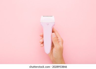 Young adult woman hand holding electric epilator on light pink table background. Pastel color. Closeup. Female product for smooth body skin.