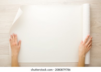 Young Adult Woman Hand Holding And Showing Opened Sheet Of Wallpaper Roll On Light Wooden Table. Closeup. Point Of View Shot. Preparation For Repair Work Of Home. 