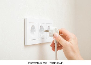 Young Adult Woman Hand Holding And Plugging White Electrical Plug In Wall Outlet Socket At Home. Closeup. Side View.