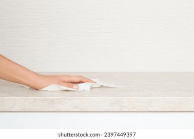 Young adult woman hand holding dry white paper napkin and wiping beige stone tabletop at home kitchen. Closeup. Cleaning service. Front view. Empty place for text on wallpaper background.