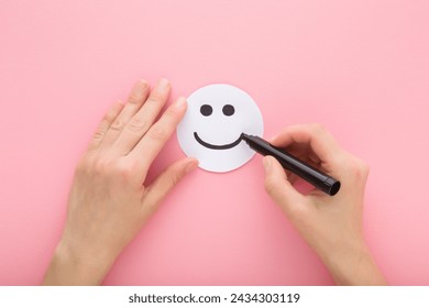 Young adult woman hand holding black marker and drawing happy smiling face on white paper on light pink table background. Pastel color. Closeup. Point of view shot. - Powered by Shutterstock