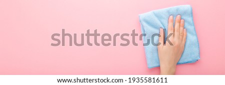 Young adult woman hand holding blue rag and wiping table, wall or floor surface in kitchen, bathroom or other room. Closeup. Light pink background. Pastel color. Wide banner. Empty place for text. 