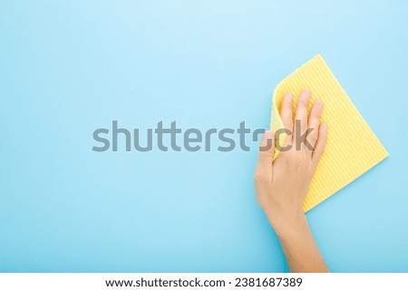 Young adult woman hand holding yellow sponge cloth and wiping table, wall or floor surface in kitchen, bathroom or other room. Closeup. Empty place for text. Light blue background. Pastel color.
