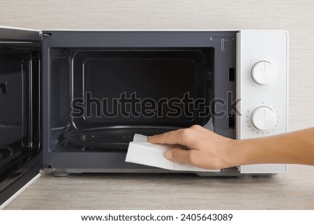 Young adult woman hand holding dry white paper napkin and wiping dark black surface of white microwave oven inside on table top at home kitchen. Closeup. Regular cleanup. Front view.
