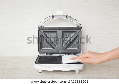 Young adult woman hand holding dry white paper napkin and wiping dark black nonstick surface inside sandwich maker on stone table top at home kitchen. Closeup. Front view.