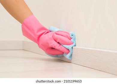 Young adult woman hand in pink rubber protective glove using blue dry rag and wiping light wooden baseboard surface in room at home. Closeup.