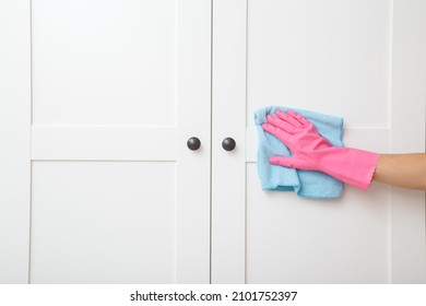 Young adult woman hand in pink rubber protective glove using blue dry rag and wiping white wooden doors of wardrobe. Closeup. Regular cleanup at home. Front view.