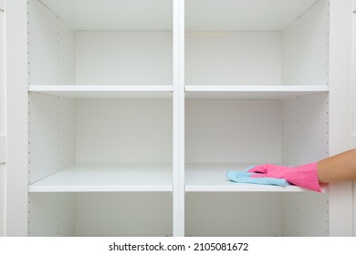 Young adult woman hand in pink rubber protective glove using blue dry rag and wiping white shelves inside wooden wardrobe in room. Closeup. Front view. Regular cleanup at home. 