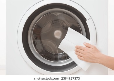 Young adult woman hand using dry paper napkin and wiping glass door surface outside white washing machine at home. Closeup. Regular cleanup. Front view. - Shutterstock ID 2394927373