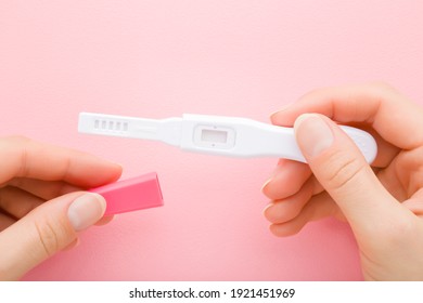 Young adult woman hands holding new pregnancy tests on light pink table background. Pastel color. Waiting negative or positive result. Closeup. Point of view shot.