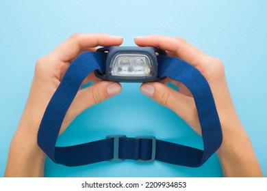Young Adult Woman Hands Holding Led Headlamp With Elastic Strap. Light Blue Table Background. Pastel Color. Closeup. Point Of View Shot. Equipment For Work Or Sport Activities In Dark Time.