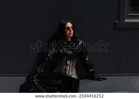 Young adult woman in latex outfit city street background. Black minimalist leather clothes at sunny day, contrast. Wearing Panama hat, sunglasses and leopard design corset. Outside photo shoot