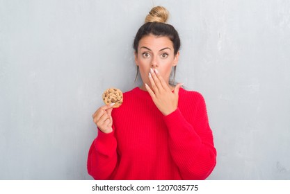 Young adult woman over grey grunge wall eating chocolate chip cooky cover mouth with hand shocked with shame for mistake, expression of fear, scared in silence, secret concept