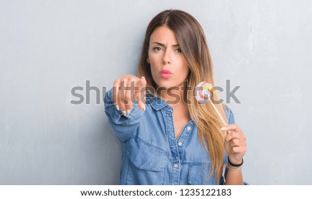 Young adult woman over grey grunge wall eating lollipop candy pointing with finger to the camera and to you, hand sign, positive and confident gesture from the front