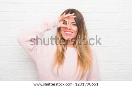 Young adult woman over white brick wall wearing winter outfit at home doing ok gesture with hand smiling, eye looking through fingers with happy face.