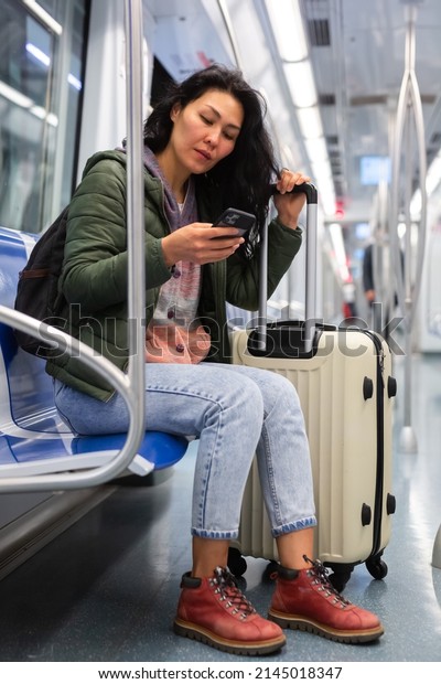 Young adult woman passenger with luggage\
sitting in subway car and using mobile\
phone