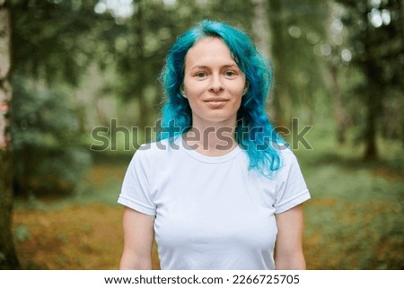 Young adult woman portrait in white T shirt with turquoise dyed hair on green forest background, attractive female artist with bright appearance. Charming woman with marine style turquoise dyed hair